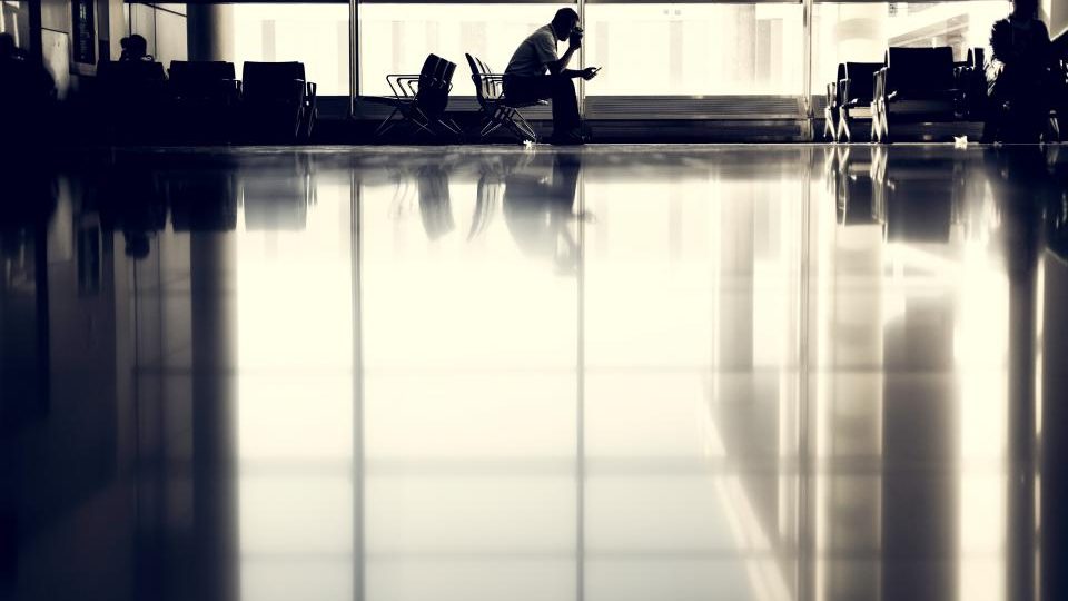 A traveller sitting alone in an airport terminal, silhouetted against large windows